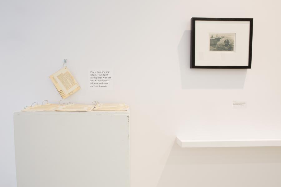 A close-up of a gallery display with archival information on small booklets, a framed photograph on the wall, and a sign explaining the display.