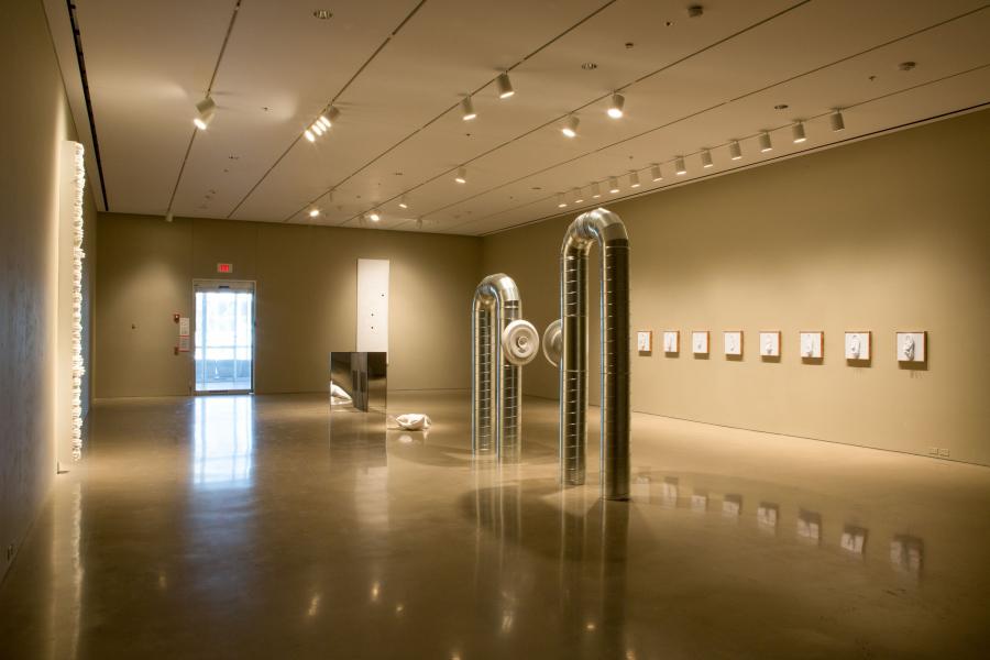 A wide shot of the gallery installation with a focus on the metallic pipe sculptures, a wall installation to the left, and the gallery entrance visible in the background.