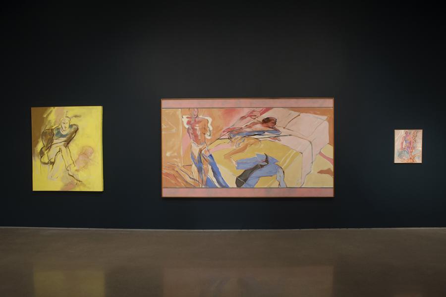 A trio of Sheila Butler’s paintings displayed against a dark wall, emphasizing their warm and energetic palette, depicting human forms and abstracted landscapes.