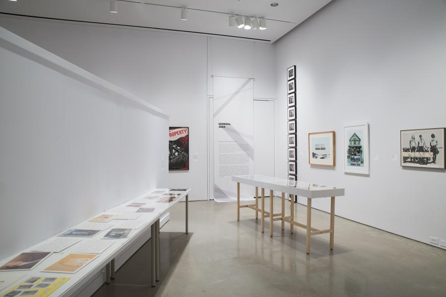 A gallery showcasing framed artworks, a "General Idea" text banner, and tables with archival documents under glass.