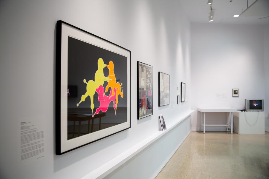A vibrant framed artwork featuring abstract, colourful figures displayed on a white gallery wall alongside other framed pieces.