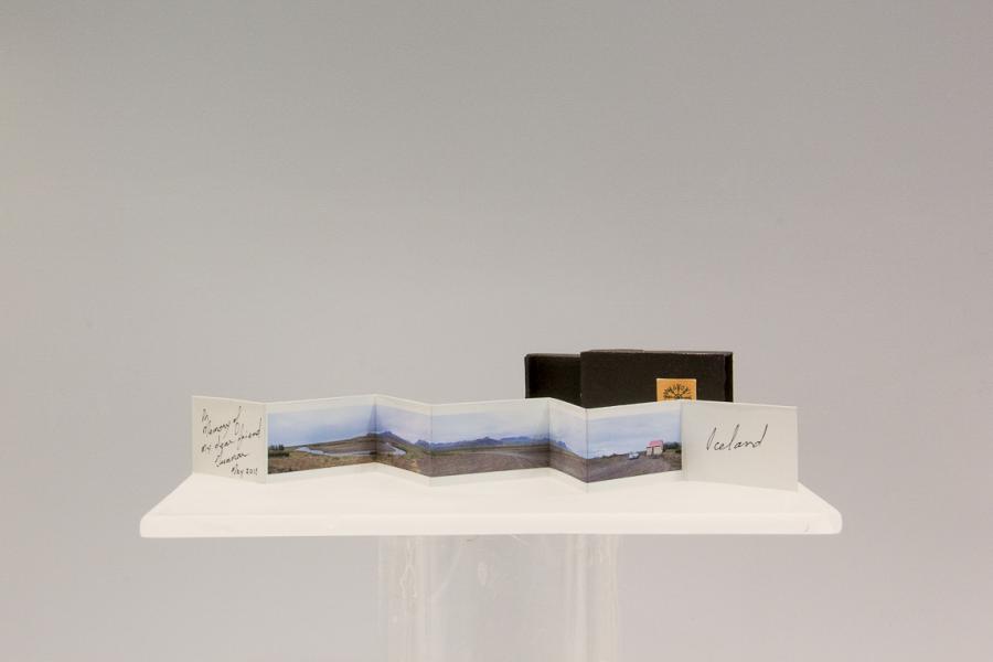 A miniature accordion book titled "Iceland" displayed on a small pedestal. The book depicts a panoramic rural landscape, with handwritten text on the cover.