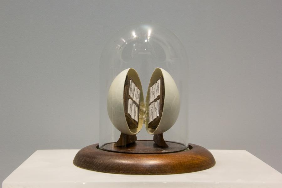 A glass-domed artwork encapsulates an open egg with book pages inside, displayed on a wooden base.