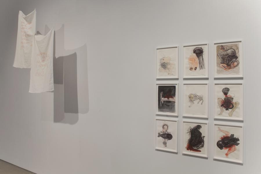Fabric pieces with embroidered text hang alongside framed drawings on a gallery wall.