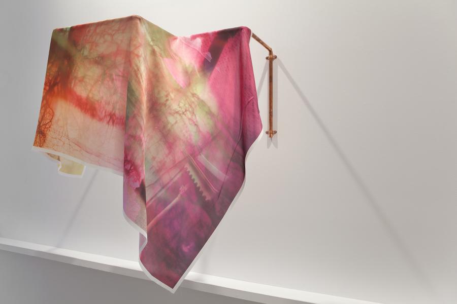 A fabric-based artwork draped over a copper pipe, displaying an abstract photographic print in hues of pink, red, and green.