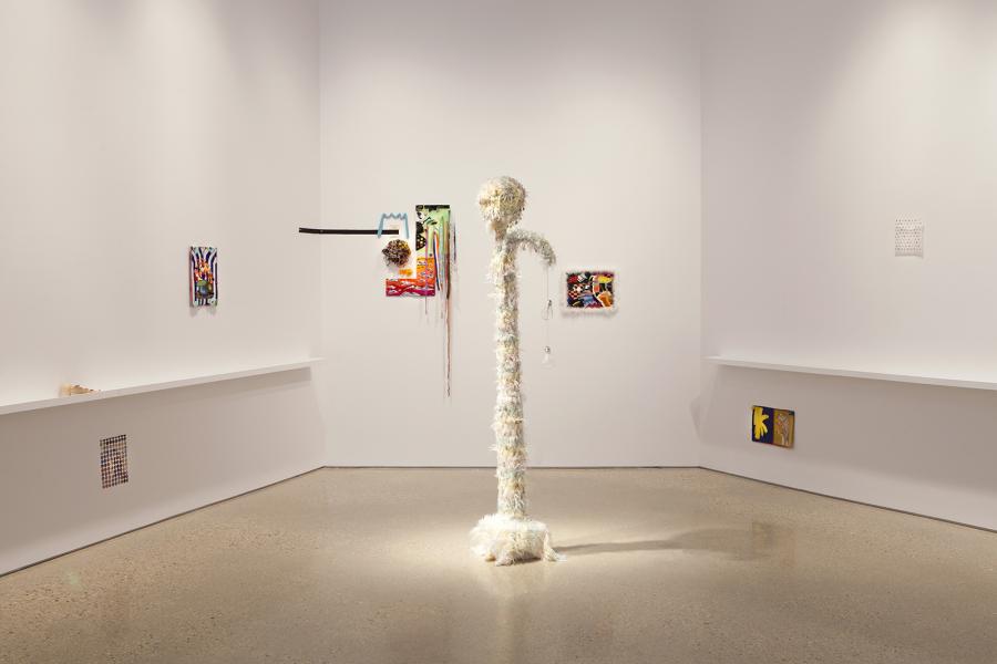 A gallery installation featuring a central tall, textured sculpture covered in white, feathery material with dangling charms. Surrounding it are various wall-mounted mixed-media textile works and assemblages.
