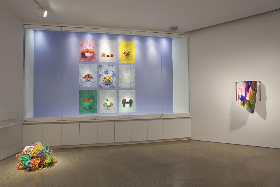 A gallery view showcasing a large backlit photographic installation of fruit bowls on the left, with a neon-colored coiled wire sculpture on the floor and a fabric piece draped over a copper rod on the right.