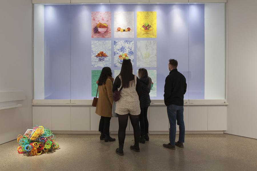 A group of four people stands in front of a backlit installation featuring nine images of fruit in decorative bowls, each set against patterned backgrounds in shades of blue, pink, yellow, and green.
