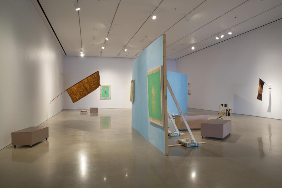 Another angle of the gallery installation, focusing on the blue partition with abstract paintings mounted on it, alongside various sculptural elements and wooden structures.