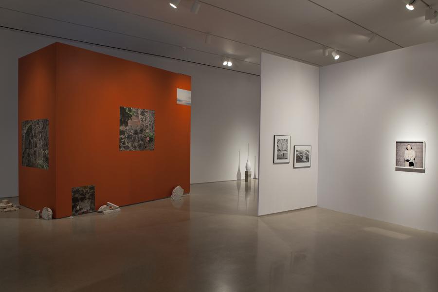 An exhibition space featuring an orange wall with embedded photographic prints, adjacent to a white wall displaying framed black-and-white images.