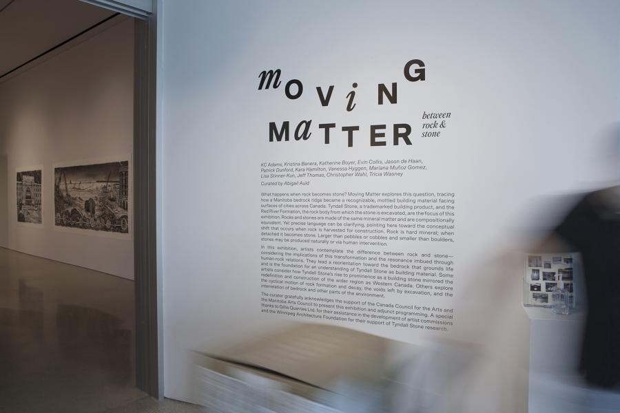 Exhibition title wall for Moving Matter, featuring bold typography and curatorial text, with a blurred figure passing by.