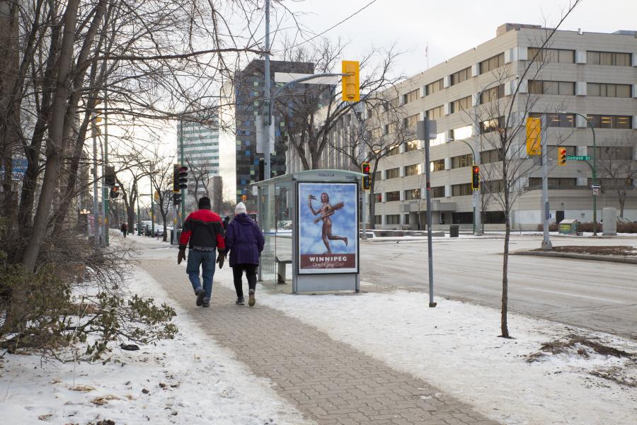 A downtown bus shelter displays an advertisement featuring a nude figure with wings, alongside the text "WINNIPEG One Gay City!" Pedestrians walk past on the snowy sidewalk.
