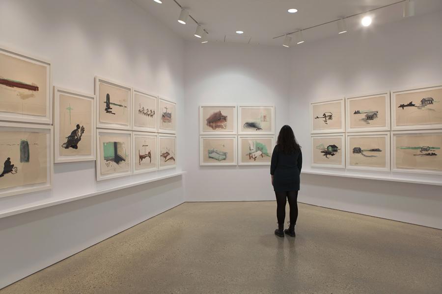 A visitor in dark clothing stands in a gallery corner surrounded by framed drawings from The Sandy Bay Residential School Series, depicting haunting scenes of beds, buildings, and figures on textured paper.