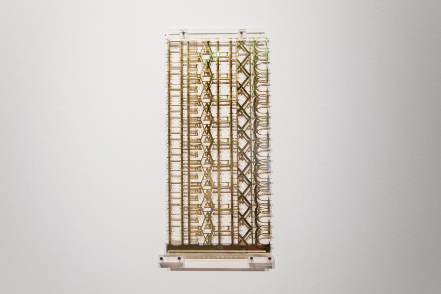 A clear acrylic wall-mounted sculpture with intricate layered patterns, resembling architectural elements. The transparency of the material interacts with the gallery lighting.