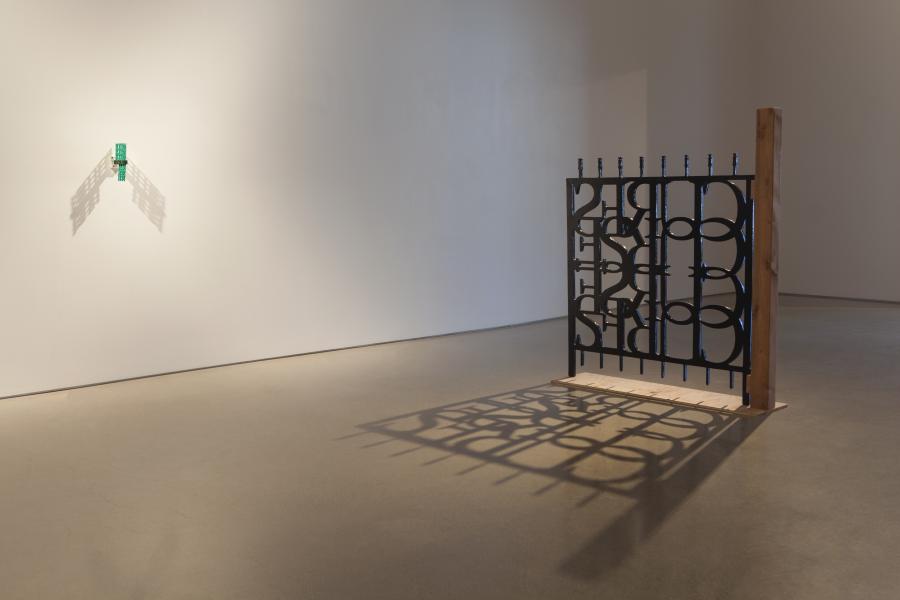 A metal gate sculpture, mounted on a wooden frame, casts a strong shadow on the gallery wall. A green bottle is placed on top of the gate.