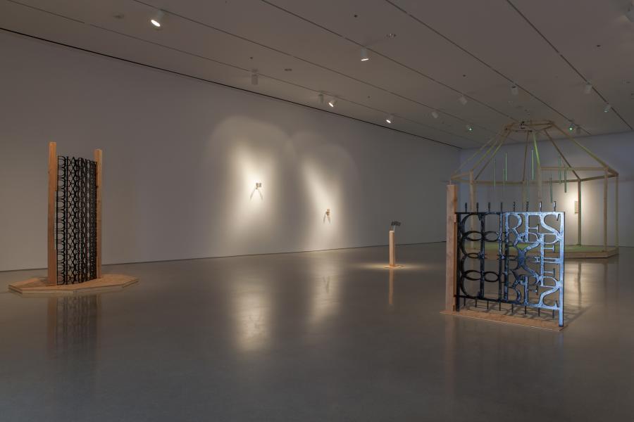 A large octagonal wooden frame resembling a gazebo stands in the gallery, with green tubes hanging from its ceiling. A small pedestal with a black metal text sculpture is positioned in front of it