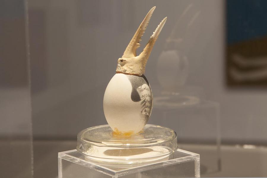 A close-up of a surrealist sculpture featuring an egg with a printed design, topped with a crab claw, displayed on a clear pedestal.