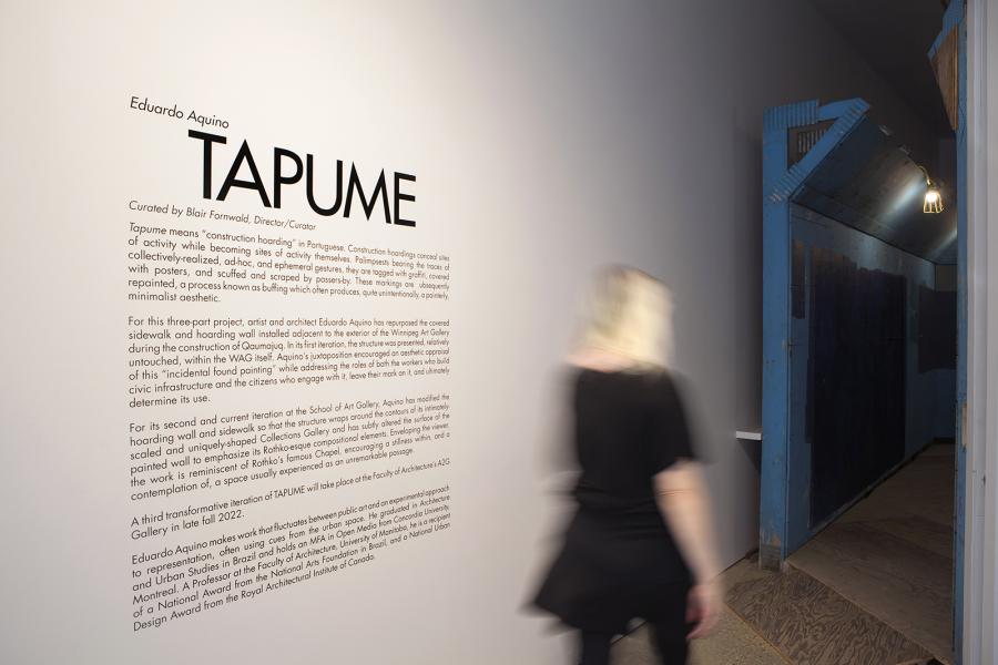 Exhibition entrance for "TAPUME," with a blurred figure walking past text detailing the installation concept.