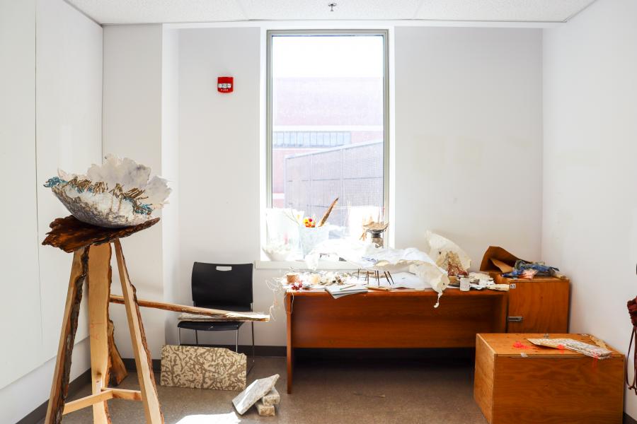 A sunlit studio space with an art installation featuring a large sculptural bowl on a wooden stand, surrounded by materials, papers, and works in progress.