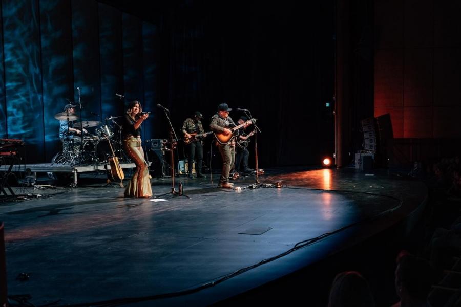 A band performs under moody blue lighting at the Desautels Concert Hall, with a singer and instrumentalists mid-performance.