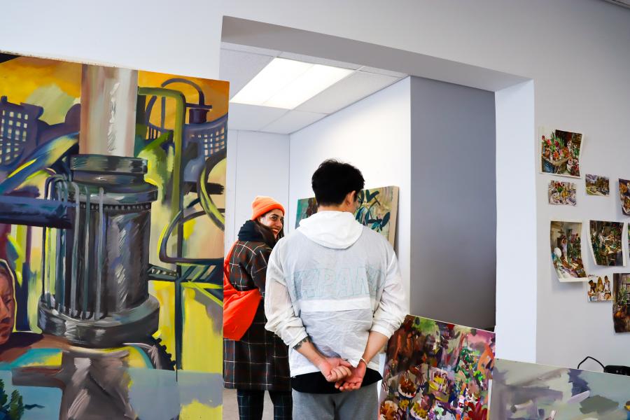 Two people talk in a studio filled with colorful large-scale paintings and collaged artworks on the walls.