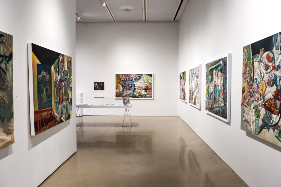 A contemporary art gallery space with white walls and polished concrete floors, displaying large-scale figurative and abstract paintings. A central table holds printed materials and a small sculpture, while vibrant, colorful canvases line both walls, depicting domestic scenes and layered compositions.