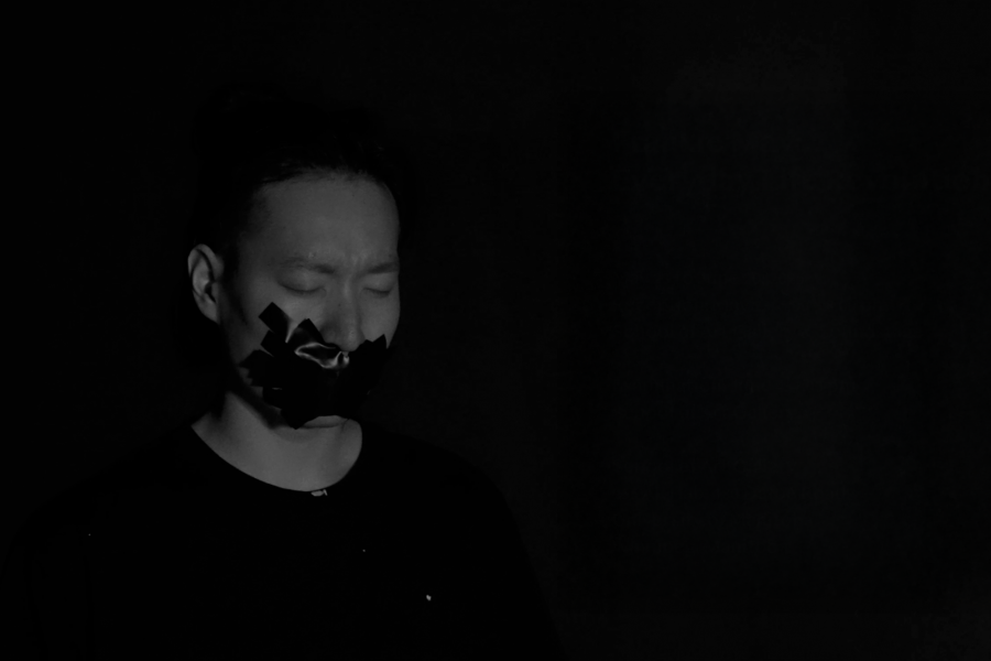 A black-and-white image of a person with their mouth taped shut, eyes closed, set against a dark background.