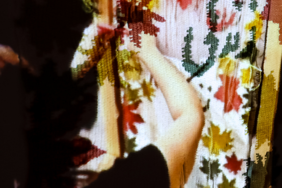 Projected image of two people engaged in artistic activity is overlaid onto a woven textile hanging in a darkened gallery space. The projection partially obscures the textile's detailed embroidery, blending figures with the fabric’s textures and patterns of colorful autumn leaves.
