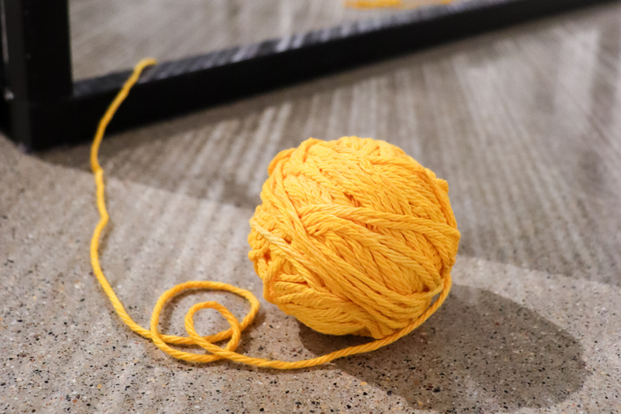 A tightly wound ball of golden-yellow yarn sits on a speckled floor. The loose end of the yarn trails outward, suggesting its connection to a larger fiber installation.