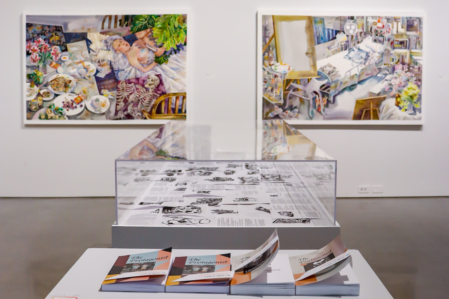 Close-up view of a gallery installation featuring two large paintings by Xianghui Guan behind a vitrine filled with printed pages, with stacks of exhibition booklets titled "The Protagonist" on a nearby table.
