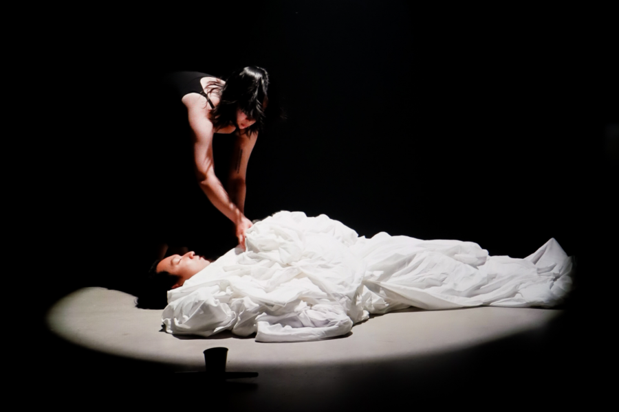 A dimly lit performance scene with one figure lying on the floor wrapped in white fabric, while another figure in a black dress leans over them gently.
