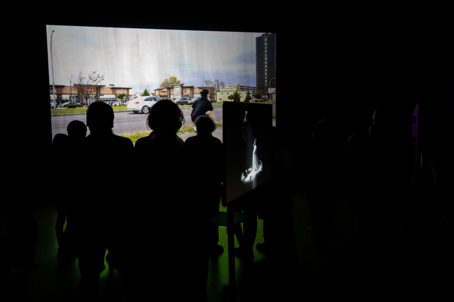 A darkened gallery with silhouetted viewers watching a video projection. The main screen shows a busy urban street; a smaller monitor displays a scene from the earlier fabric performance.