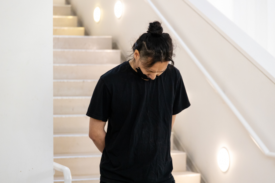 A figure in black stands on a staircase in soft lighting, looking downward. Their hands are clasped behind their back.