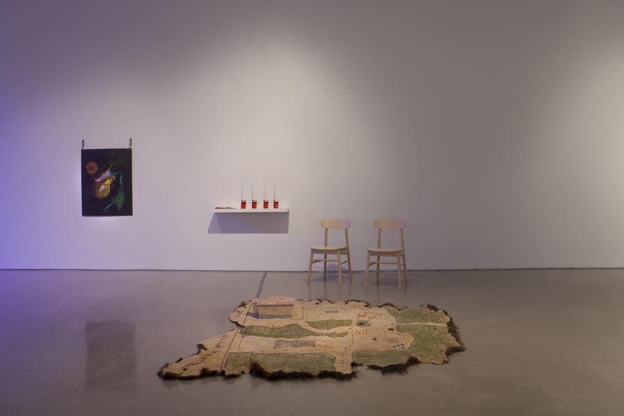 Installation view featuring a textile map laid on the floor, two wooden chairs, a shelf with four red walkie-talkies, and a hanging artwork under purple light.