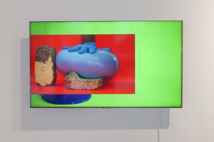 A digital video work by Elizabeth Milton displayed on a wall-mounted screen at the School of Art Gallery, University of Manitoba. The screen shows a vibrant composition with a bright green border framing a red background. At the center, a blue inflated form is pressed down by a gloved hand, resting on sponge-like materials.