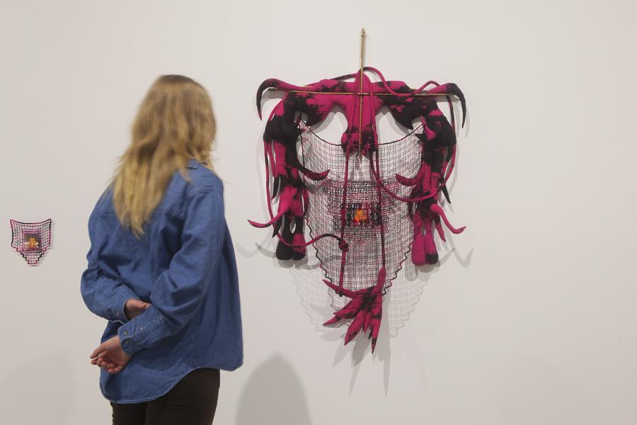 A visitor stands close to a mask-like textile sculpture made of pink-and-black fabric with protruding tendrils, stretched across a woven grid with an image embedded in the center.