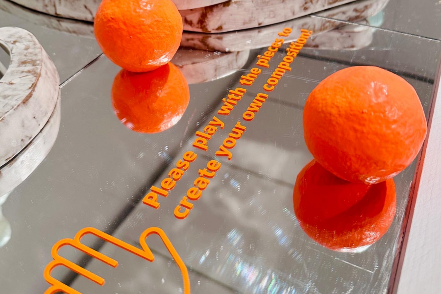 Close-up of mirrored surface with orange sculptural balls and text inviting visitors to play.