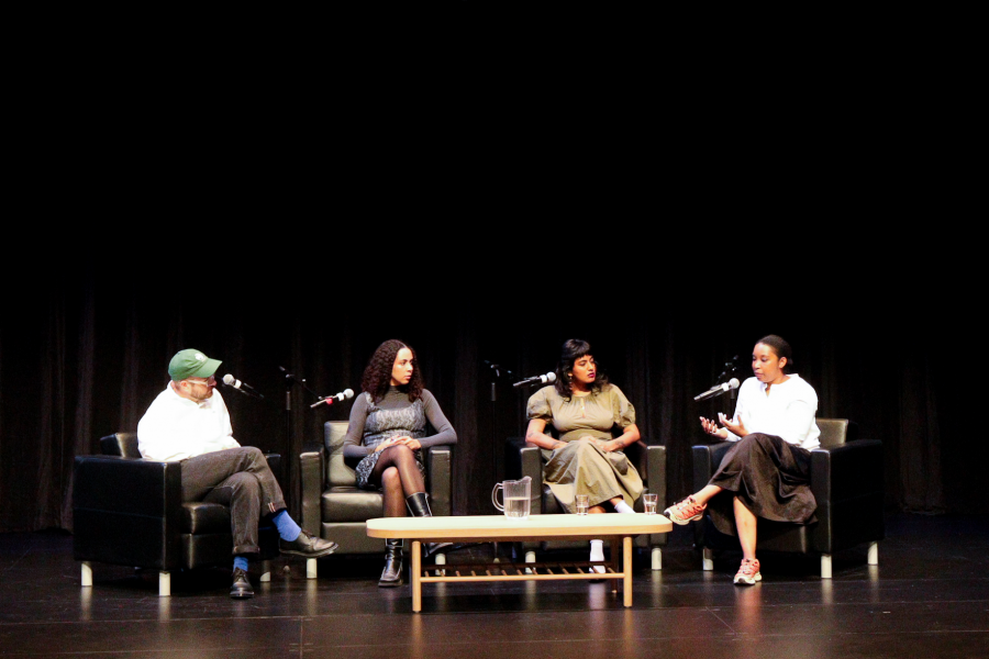 A four-person panel seated onstage in armchairs, speaking into microphones. A large projected image of an outstretched arm is displayed above them.