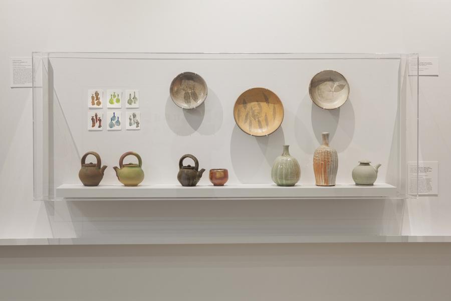 A display case shows ceramic teapots, bowls, and vessels alongside small watercolor studies.