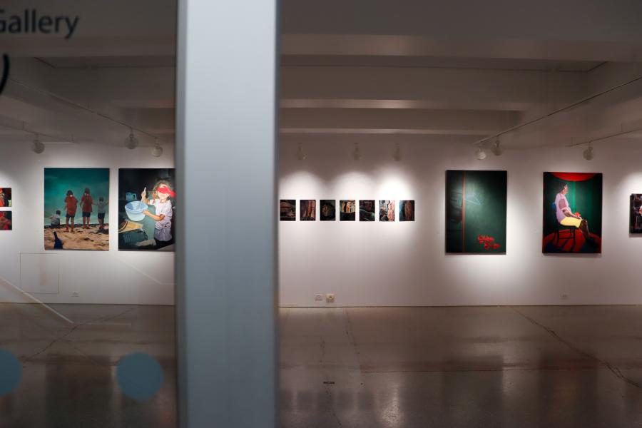 View through a glass wall into a gallery showing framed photographs and paintings arranged along white walls.