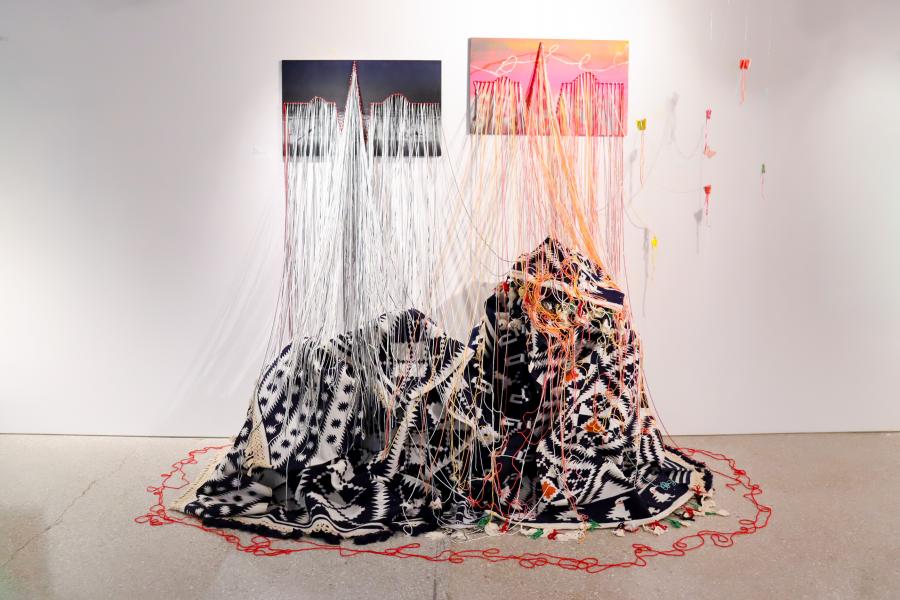 Two draped textile forms covered in hanging threads sit beneath small paintings on a gallery wall.