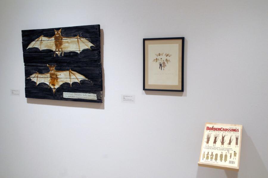 Two framed artworks depicting brown bats and a magazine on a wall. On the left is a painting of two brown bats on black on wooden boards, captioned “Common European bats can’t bite your neck, but they can steal your heart” in the lower right corner. In the centre is an illustration of brown bats attacking a man and a woman, with a small illegible caption. In the lower right is a shelf holding the magazine “drawing a fine line”, with a cover illustration depicting a tree evolving into a human.