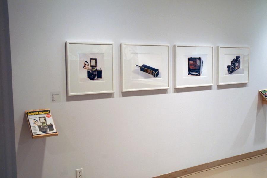Four framed photos depicting black, yellow-trimmed tech devices and a matching magazine on a wall. From left to right, the framed works depict a camera with a flash and small strap attached, a dynatac phone, a CRT television, and a camcorder. To the lower left, a shelf holds a magazine with the same camera on its cover.