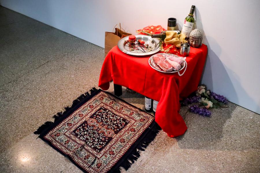 Small table draped in red fabric holds ritual objects, food, and bottles beside a patterned rug on the gallery floor.
