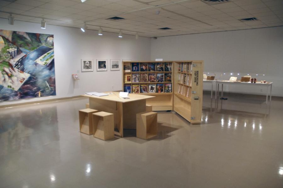 The gallery with a wooden table, stools, and magazine rack in the centre. A large blue and green painting is partly visible on the left side. A set of framed black and white images and a display table with brown items are both partly visible behind the magazine rack.