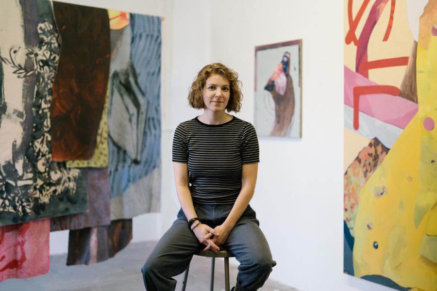 Portrait of M.E. Sparks seated on a stool in a gallery space, with large, layered textile artworks hanging behind her.