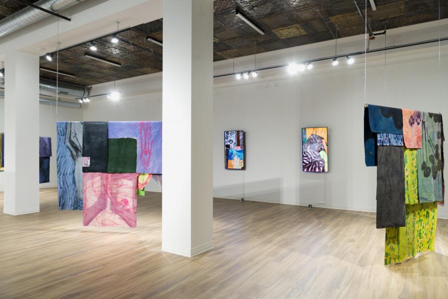 Wide gallery view showing multiple hanging textile works and framed pieces, emphasizing depth, colour, and spatial rhythm.