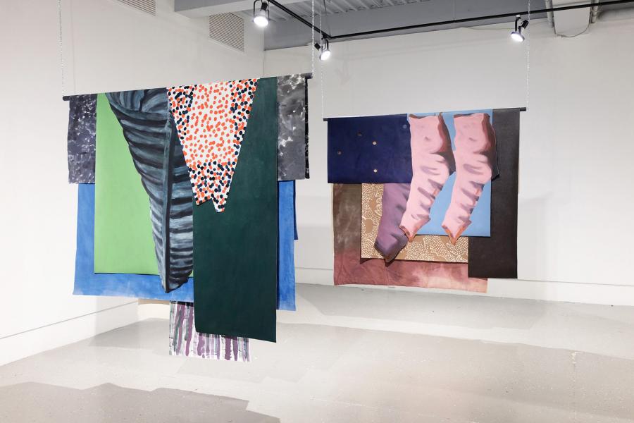 Close view of two suspended textile works with abstract imagery and layered fabrics, hanging parallel in a white gallery space.