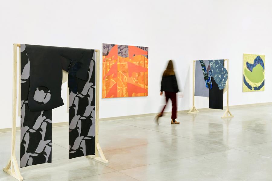 Gallery view showing freestanding wooden supports holding painted textile panels, with framed paintings visible along the walls.