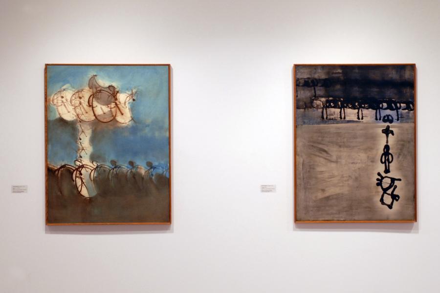 Two abstract paintings on a wall. The left one is blue on top and grey underneath, with a white T shape and black circular lines on it. The right one is grey at the bottom and black overtop, with a blue line in between and black circular lines on it, and black line symbols stacked vertically in the lower right corner.